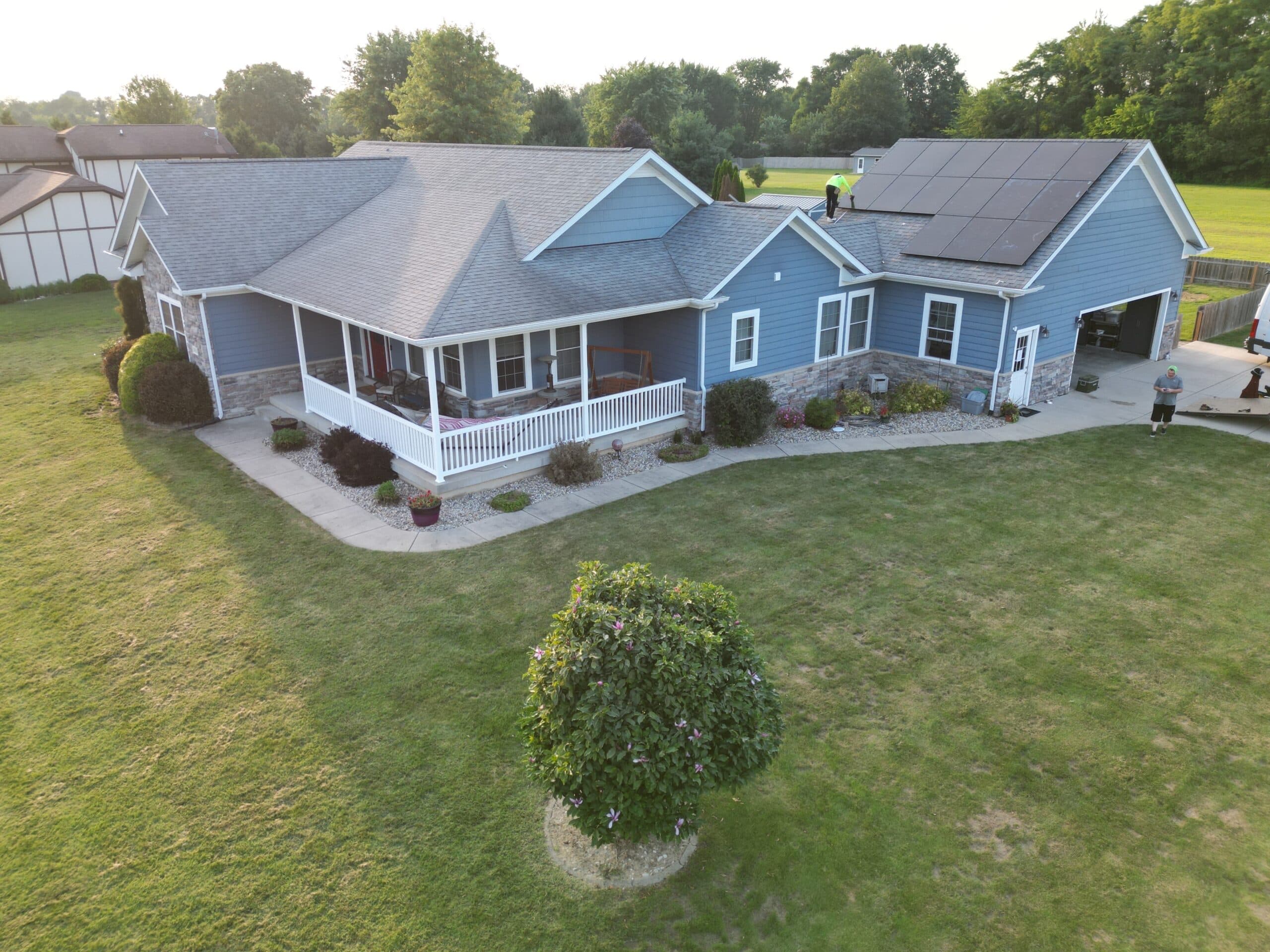 Aerial drone view of blue ranch-style home with solar panels being installed by crew on the roof